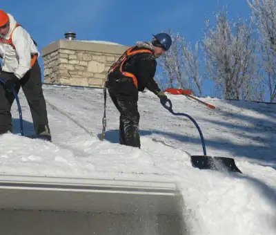 Professional technicians performing roof snow removal with safety harnesses and shovels in Rochester, NY